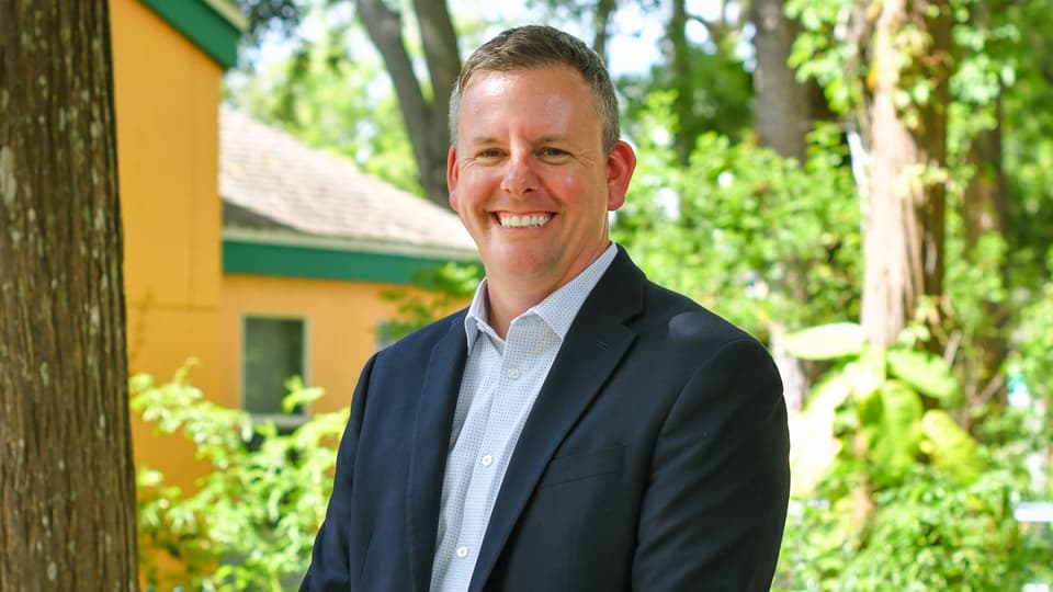  A man looks into the camera and smiles. In the background are bushes and part of a yellow house.