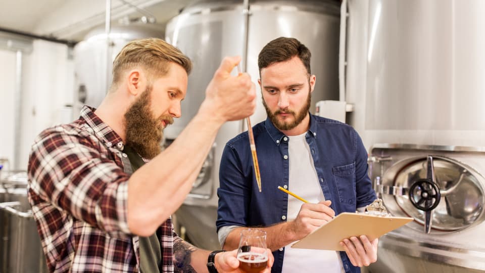 Två män testar öl i ett mikrobryggeri. En man håller i ett glas med öl och den andra antecknar. I bakgrunden syns öltankar. 