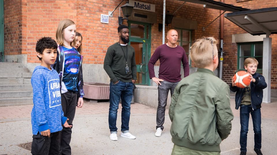 Barn leker med boll på skolgård, två vuxna närvarande.