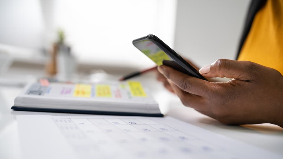 En hand håller i en mobiltelefon och en kalender ligger på ett bord.
