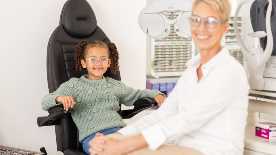 Ett barn med glasögon sitter i en optikerstol. En kvinna sitter framför henne. Båda ler.