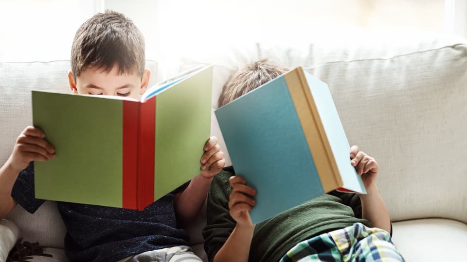Two boys are sitting on a sofa reading books.