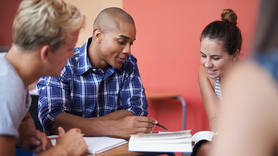 Group of four students in a classroom. In front of them is a book that they seem to be discussing. Behind them there is a red wall.