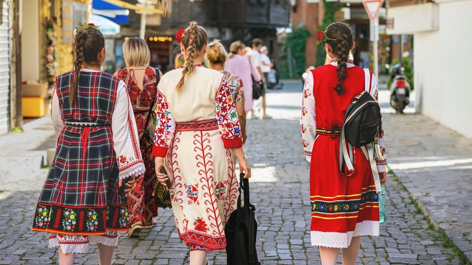 Tre kvinnor i traditionella bulgariska folkdräkter går på en gata i en stad. Dräkterna är färglada med starka färger och mönster. 