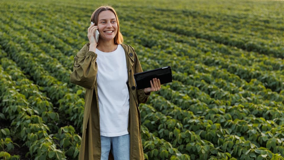 Young female farmer standing in a big field where beans grows. She is holding a phone in one hand and an ipad in the other hand. She looks happy.  
