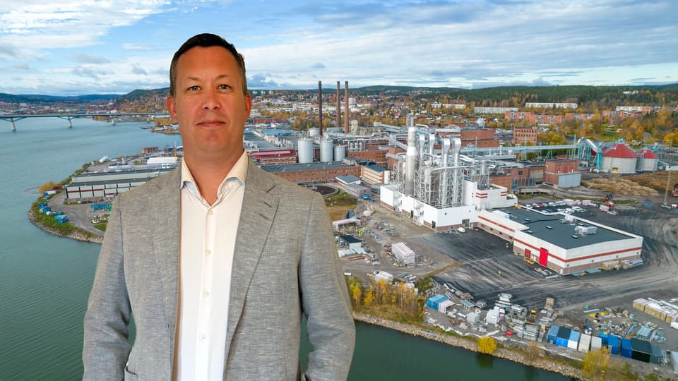 En man står framför en bild av ett fabriksområde.