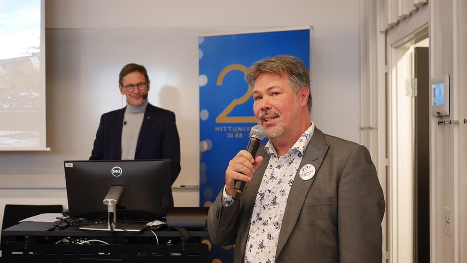 En man i grå kavaj och mönstrad skjorta står bredvid ett podium och håller i en mikrofon.
