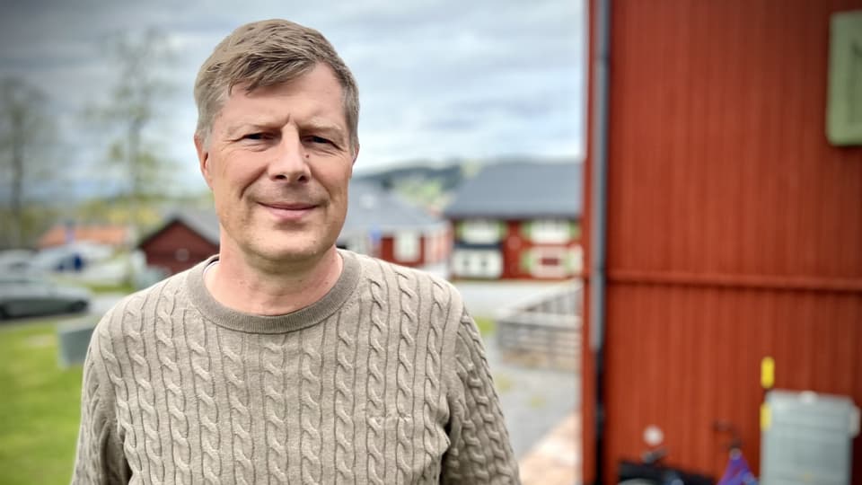 Man i beige tröja står utomhus med en röd husvägg i bakgrunden. 
