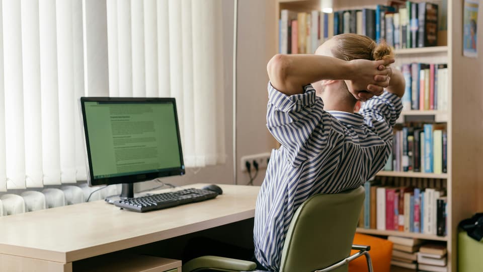 Person lutar sig lite bakåt med händerna bakom huvudet vid ett skrivbord med en datorskärm