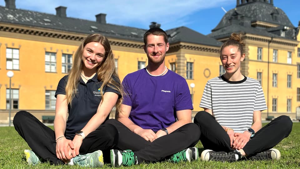 Tre unga människor sitter på gräs framför gul byggnad.