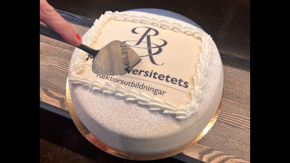 En vit marsipantårta med texten Mittuniversitetets rektorsutbildningar och en hand som håller i en tårtspade och precis ska skära i tårtan.