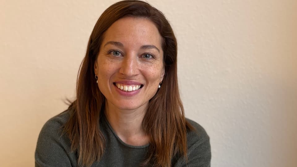 Kvinna med långt brunt hår med fräknar, ler mot kameran, klädd i grå tröja.