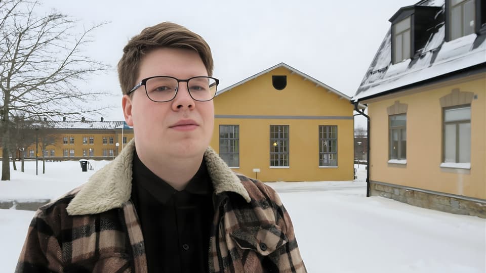 Manlig student står på campusgården i Östersund framför några av byggnaderna. Gården är snötäckt och studenten har vinterjacka på sig.