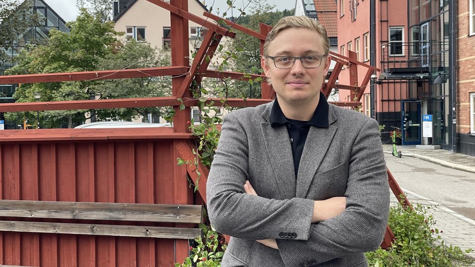 En ljushårig man med glasögon och kavaj står framför ett rött staket och poserar framför kameran.