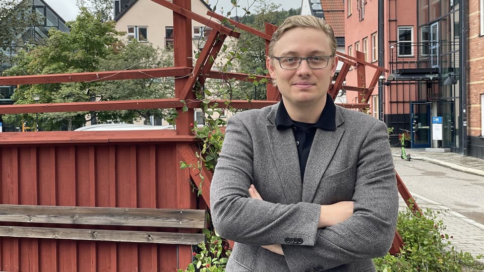 En ljushårig man med glasögon och kavaj står framför ett rött staket och poserar framför kameran.