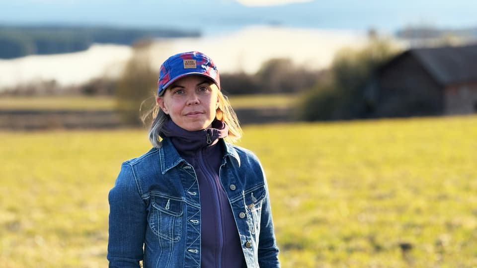 En kvinna i jeansjacka och keps står på en äng. I bakgrunden syns en sjö och fjäll.