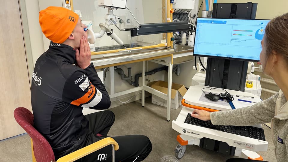 Ung man med toppluva blåser i ett munstycke, testledare övervakar på bildskärm.