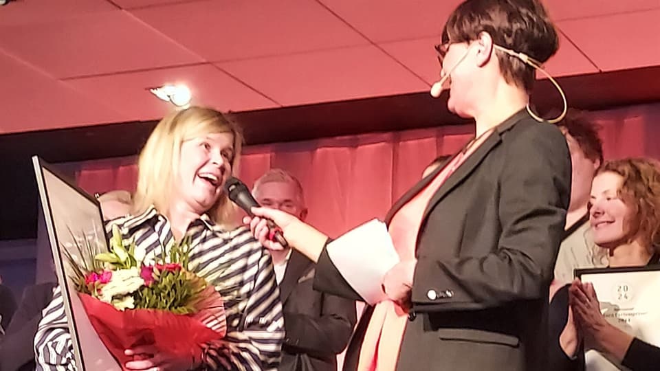 Två kvinnor står på en scen med människor bakom sig som apploderar.. Den ena håller fram en mikrofon åt den andra som står med ett diplom och blommor i famnen. 