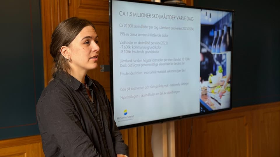En kvinna i brunt hår och mörkgrå skjorta står vid en skärm med en presentation på. 