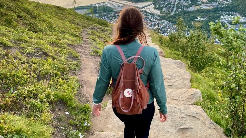 En kvinna med ryggsäck på ryggen går på en stenbelagd stig och ser ut över en norsk fjord.