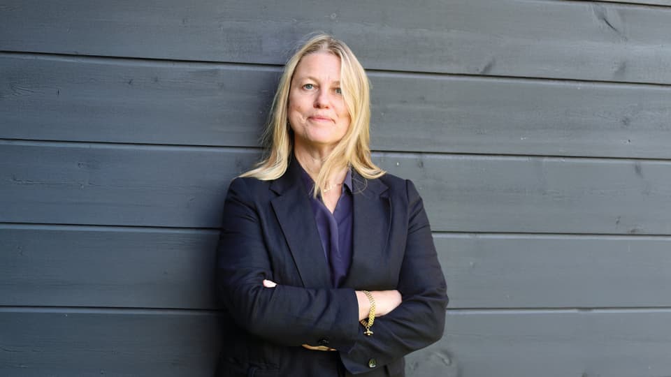 En blond kvinna står med armarna i kors framför en svart trävägg.