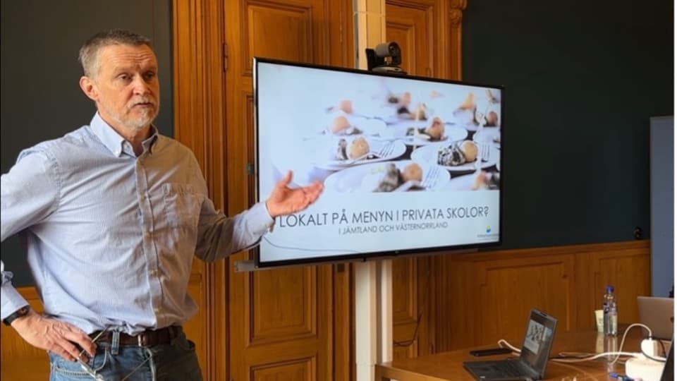 En man i ljusblå skjorta står framför en skärm med en presentation på och gestikulerar med sin vänstra hand.