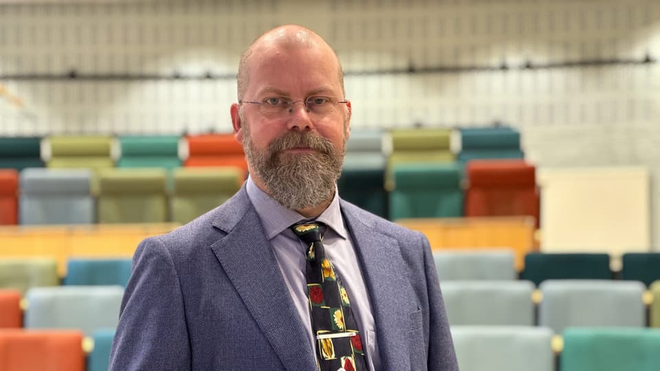 En man i blå kavaj visar upp sin doktorsavhandling i en sal med fägglada stolar