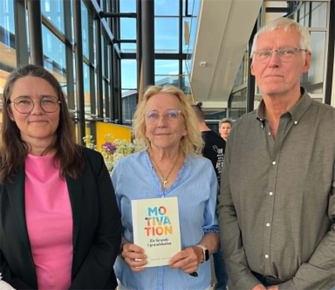 Två kvinnor och en man ler mot kameran och håller upp en bok