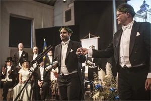 Two well-dressed people wearing laurel wreaths are standing on a stage.