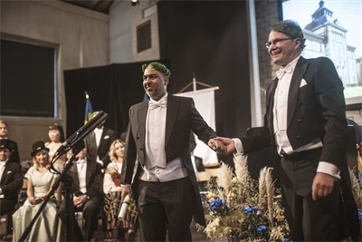 Two well-dressed people wearing laurel wreaths are standing on a stage.