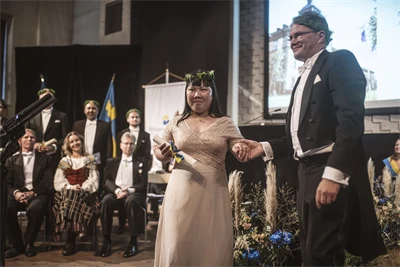 Two well-dressed people wearing laurel wreaths are standing on a stage.