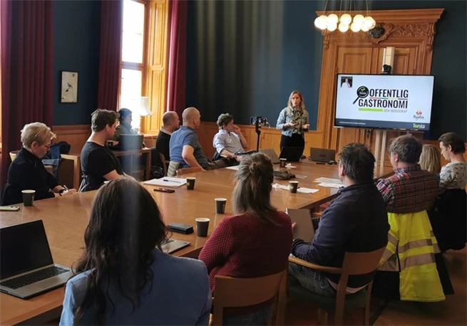 Ett toital personer i ett rum där en kvinna står längst fram och presenterar framför en skärm
