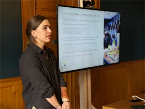 A woman with brown hair and a dark grey shirt stands by a screen with a presentation on it.